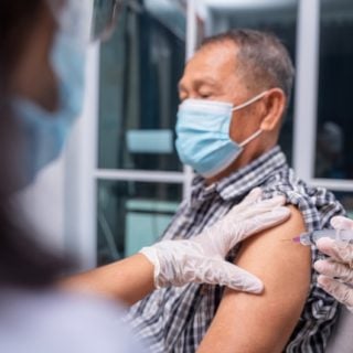 A man with grey hair is wearing a mask and receiving a vaccine injection in his left arm